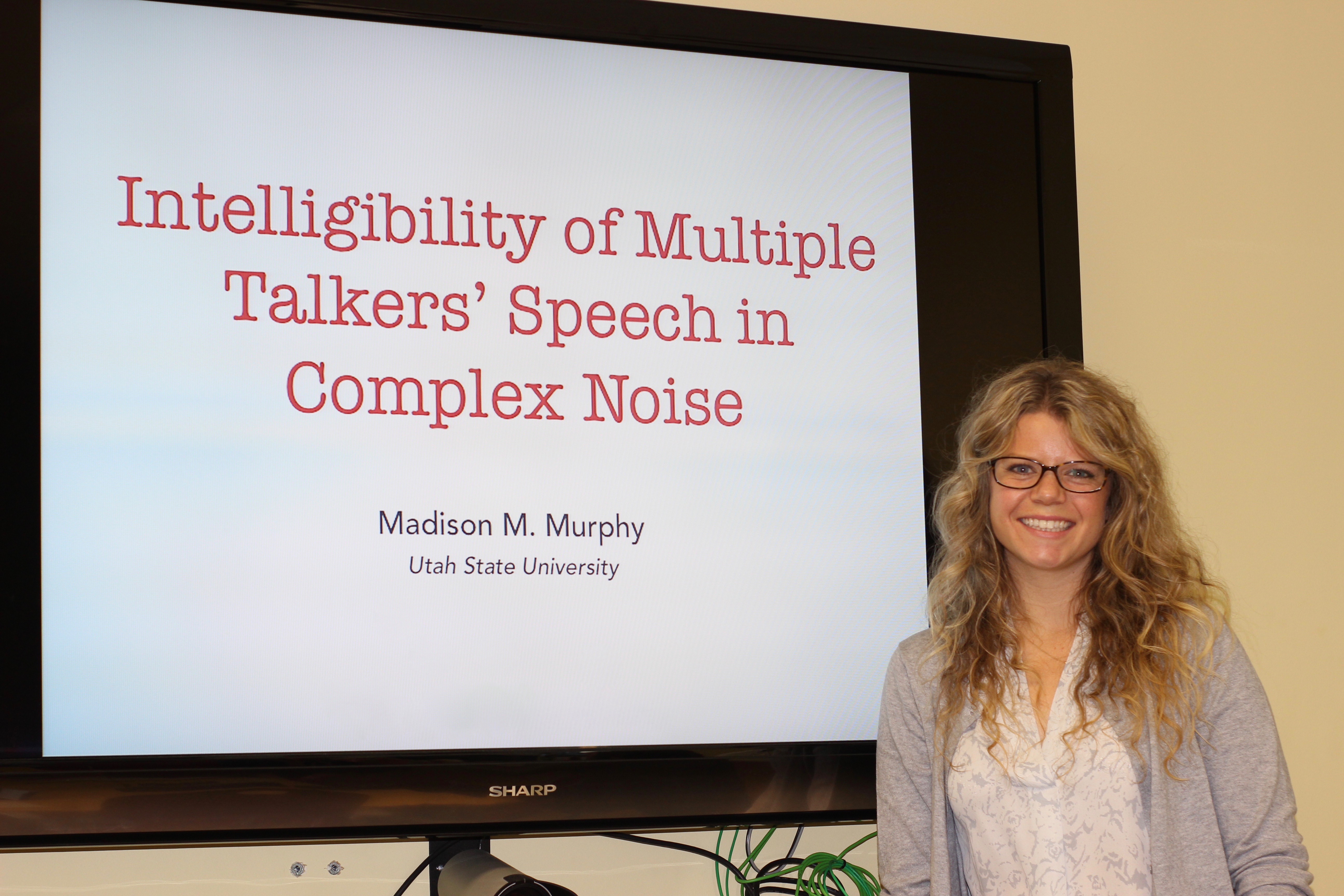 white, woman with glasses and long blonde hair standing in front of a large monitor that reads, "Intelligibility of multiple talkers' speech in complex noise. Madison M. Murphy, Utah State University".